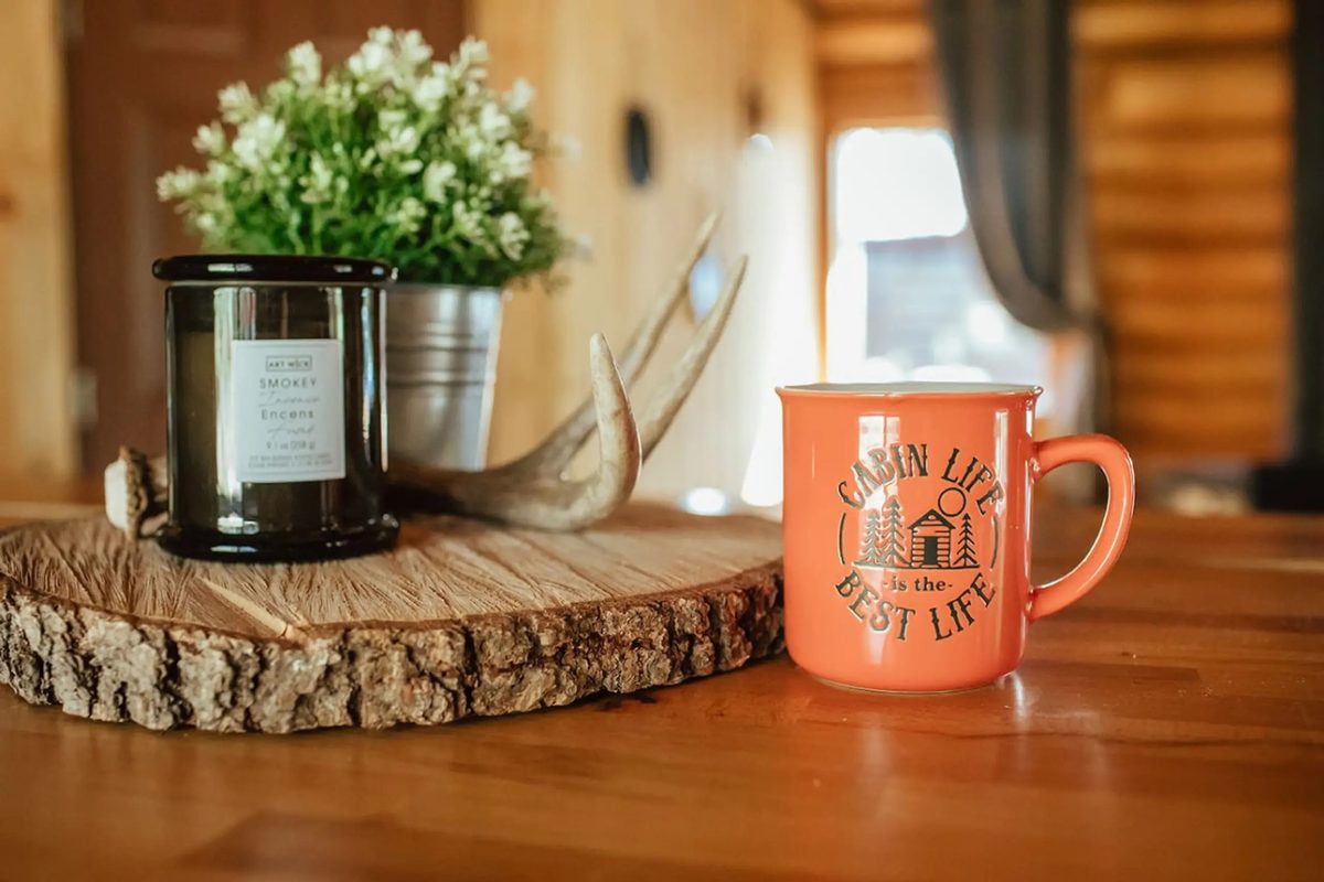 A “cabin life is the best life” mug beside a candle and greenery on a live-edge cabin table